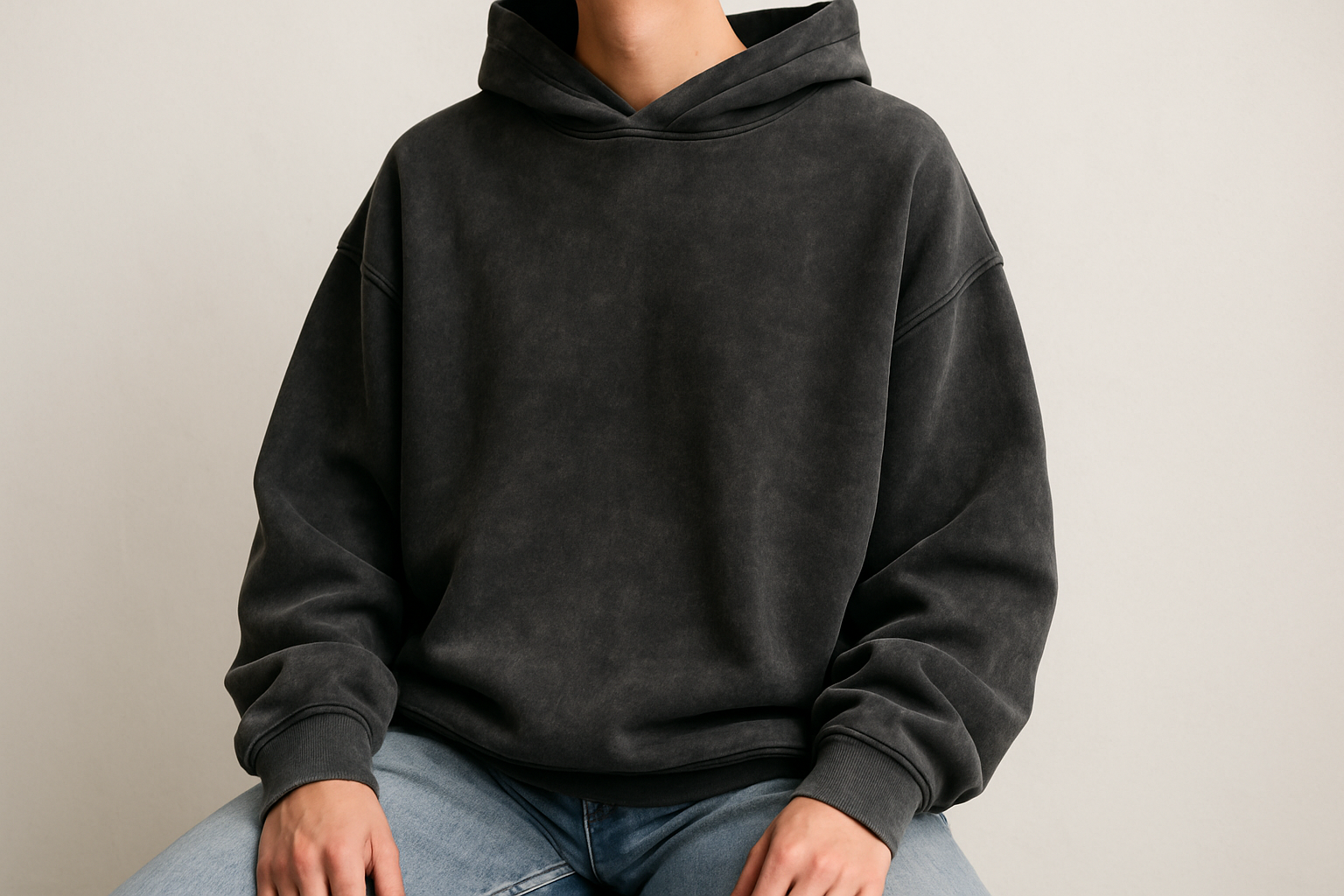 schwarzes washed out oversize hoodie ohne tasche ohne Kordeln getragen von einer Person von der front von der man das Gesicht nicht sieht, locker sitzend baumwolle, Heller hintergrund